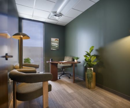 A modern office with green walls, a wooden desk, swivel chair, two upholstered visitor chairs, a floor lamp, potted plants, and artwork on the wall, with natural light coming through a tall window.