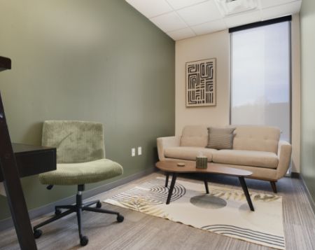 A small modern office features a green accent wall, beige sofa with pillows, wooden coffee table, green desk chair, abstract rug, and framed wall art. A large window lets in natural light.