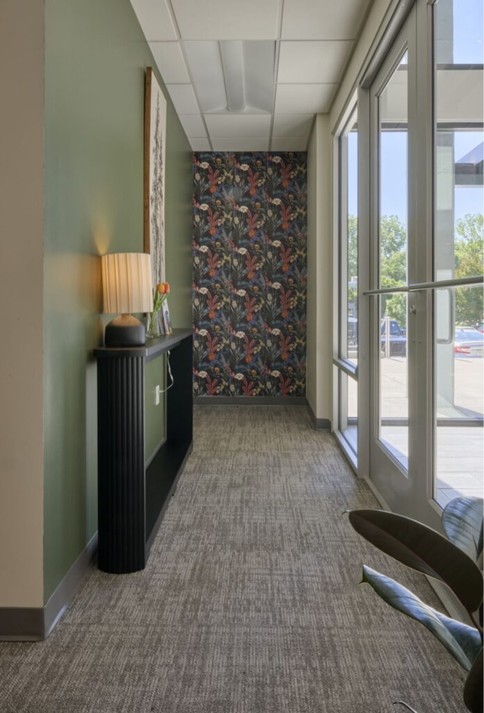 A hallway with green walls, a patterned accent wall at the end, large windows on the right, a black console table with a lamp and vase, and grey carpet flooring. Sunlight streams in through the windows.