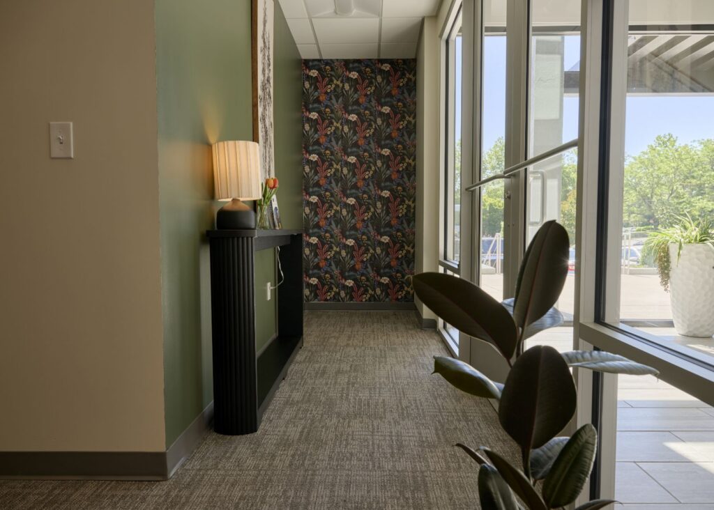 A hallway with large windows on the right, a plant in the foreground, a console table with a lamp and small vase, and a dark floral accent wall at the end. Natural light fills the space.