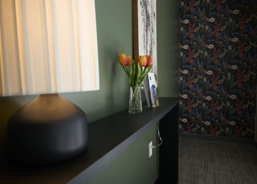 A modern black table against a green wall holds a lamp, a glass vase with orange tulips, and a framed photo. The background features floral wallpaper and gray carpeted flooring.