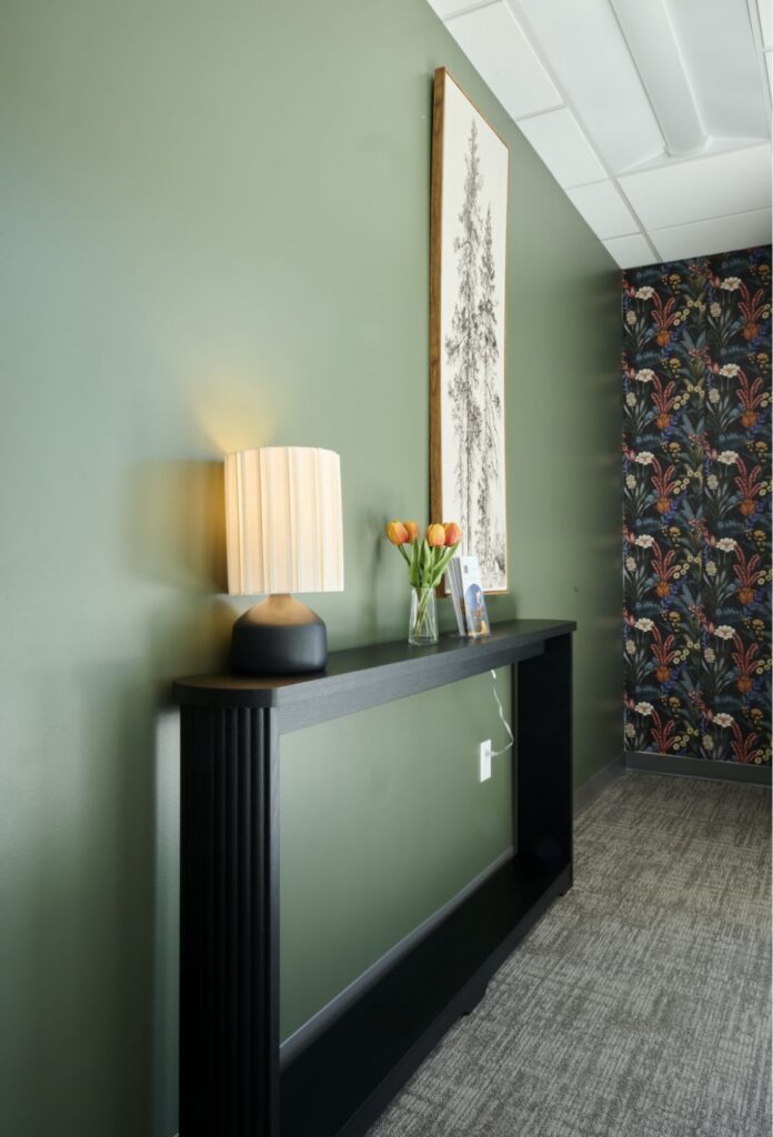 A modern interior with a green wall, black console table, lit lamp, vase of orange tulips, a framed photo, and wall art. Floral-patterned wallpaper is visible on the adjacent wall.