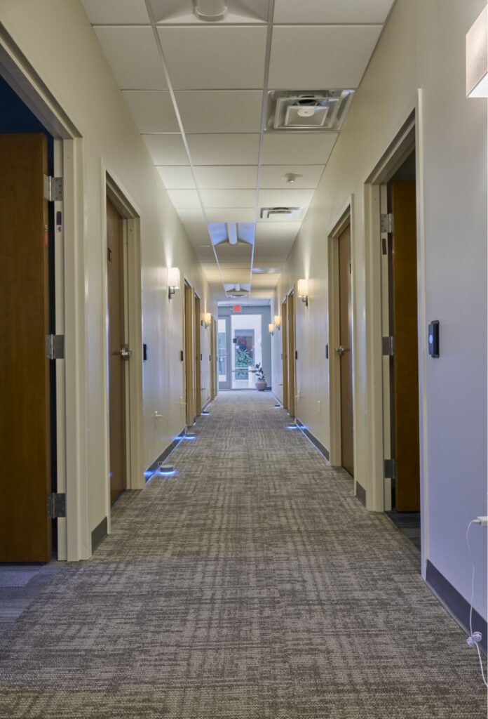 A well-lit hallway with beige walls and gray carpet, lined with open wooden doors on both sides, leads to a glass exit at the far end, where some greenery is visible outside.