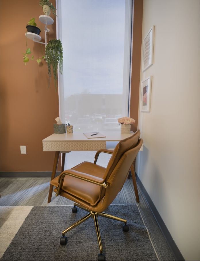 A modern office corner with a brown leather chair, a white desk holding a notebook, pen, tissue box, and flowers. A window with sheer curtains lets in daylight, and a hanging plant is above the desk.