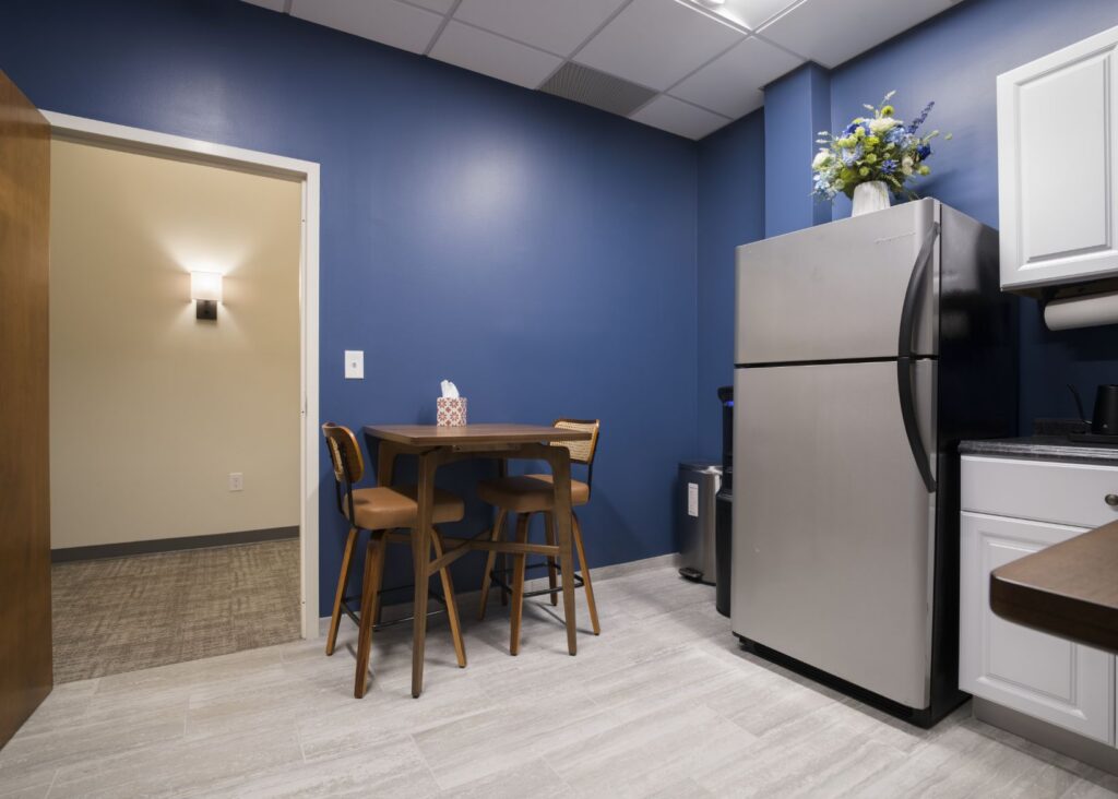 A small kitchen with blue walls, a stainless steel refrigerator, white cabinets, a wooden table with three chairs, a tissue box on the table, and a flower arrangement on top of the fridge.
