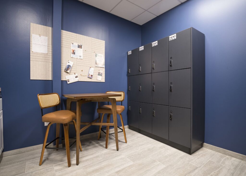 A small break room with blue walls, a wooden table and three chairs, a bulletin board with papers and photos, and a set of black lockers on a light wood floor.