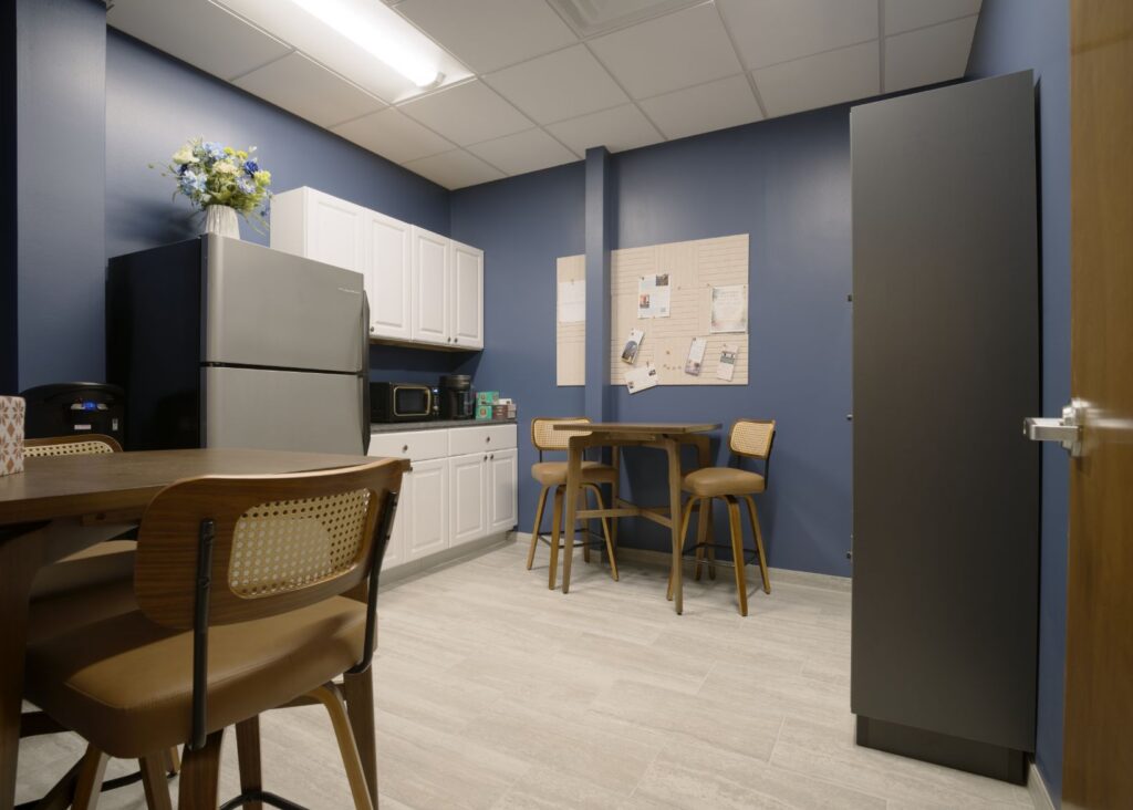 A break room with blue walls, a kitchenette with a fridge, microwave, cabinets, a small round table with two chairs, a corkboard with papers, and a wooden table with chairs. A vase of flowers sits on the fridge.
