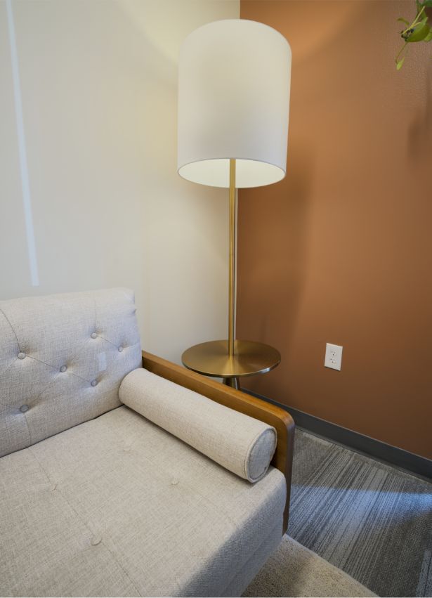 A modern beige armchair with a cylindrical pillow sits next to a round side table and a tall floor lamp with a white shade, against a two-toned wall in a cozy, well-lit corner.