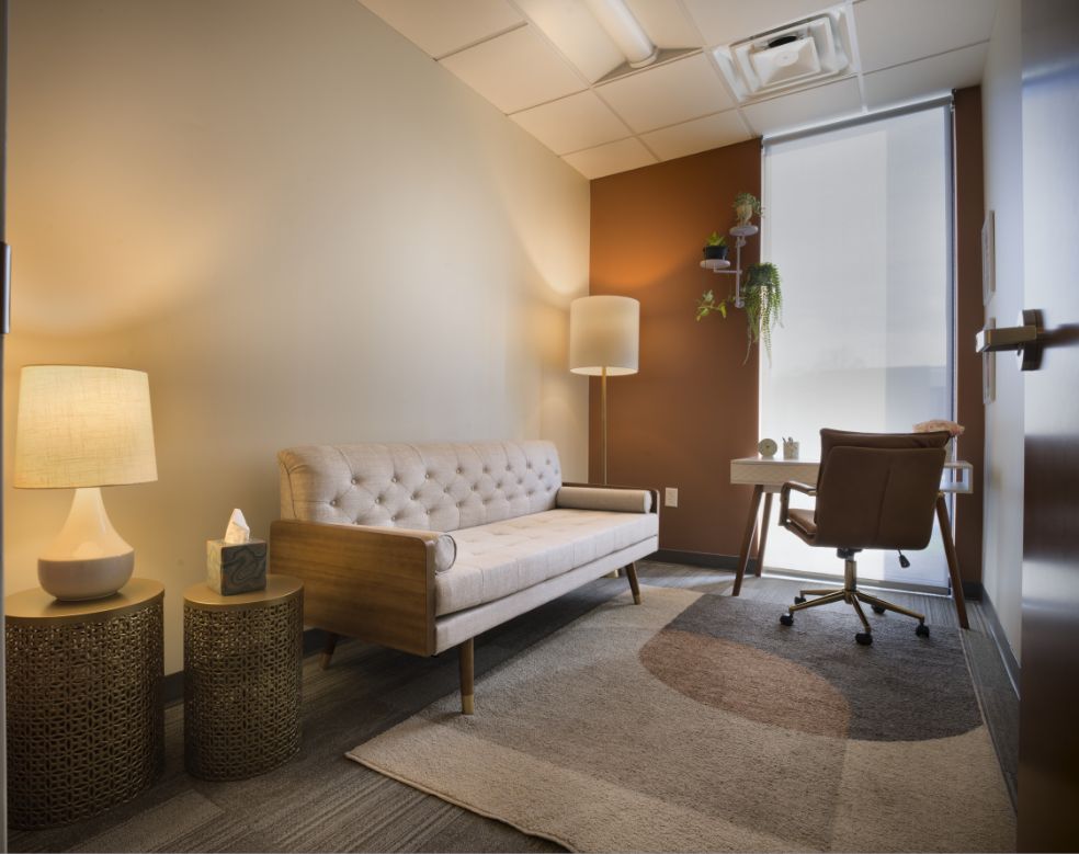 A cozy office with a beige tufted sofa, two round side tables with lamps, a floor lamp, a rug, and a small desk with a chair by a window. There are plants on shelves above the desk.