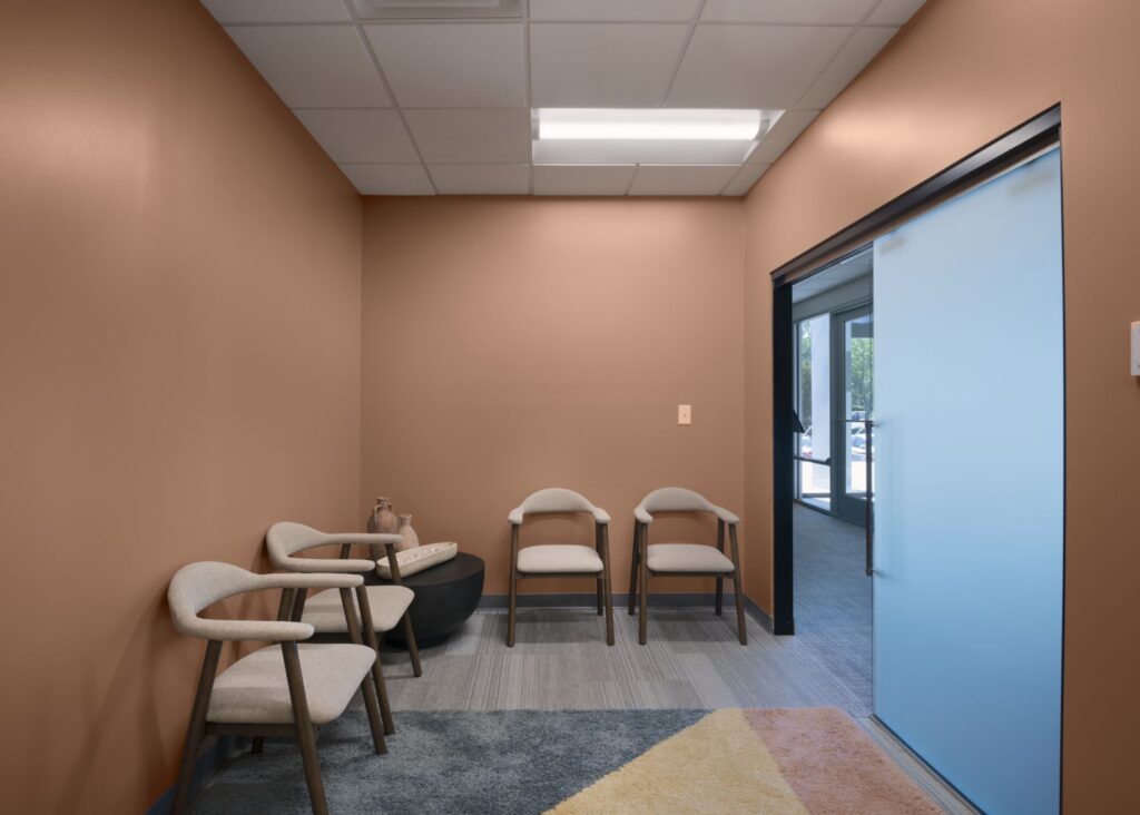 A small waiting room with three light-colored chairs, a round black table holding decorative vases, brown walls, a gray-blue rug, and a large sliding frosted glass door leading to a hallway with windows.