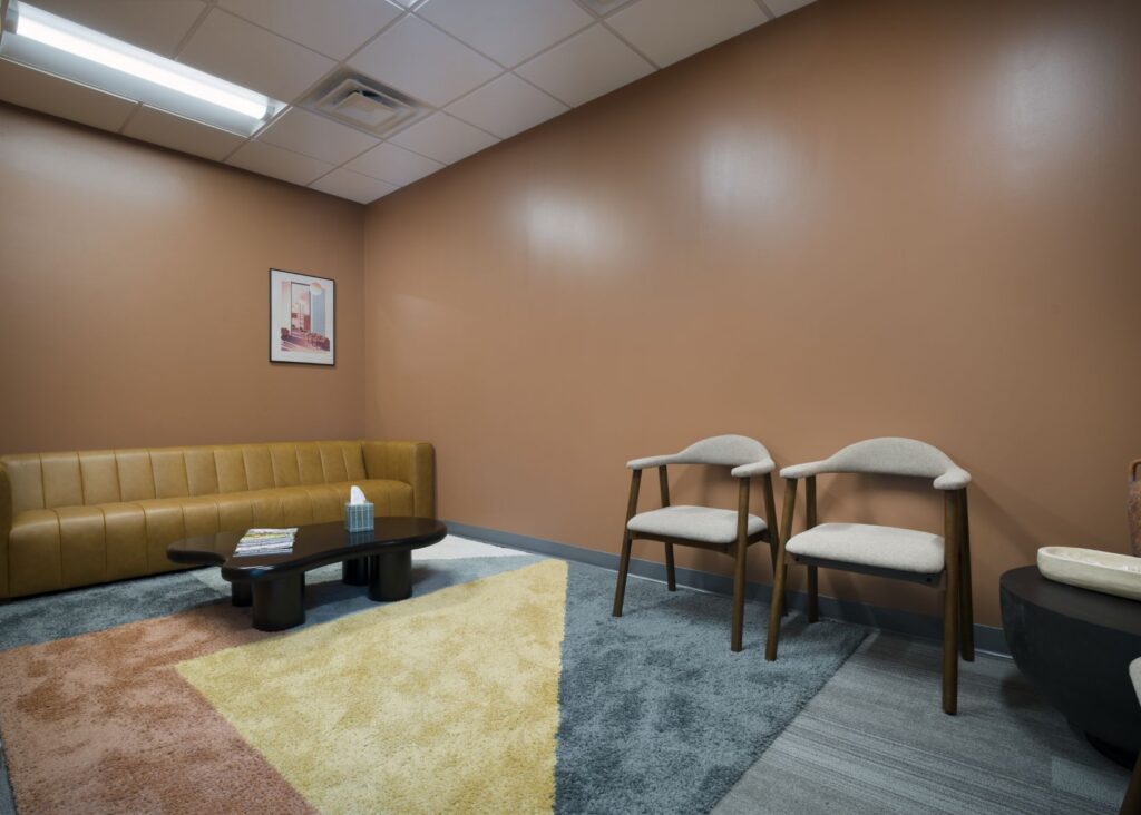 A modern waiting room with a tan couch, two beige chairs, a low black coffee table with magazines and a tissue box, a geometric-patterned rug, brown walls, and a framed picture on the wall.