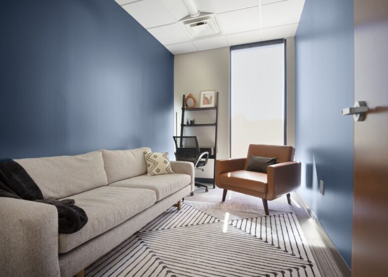 A modern office with blue walls features a beige sofa, a brown armchair, a black shelving unit with decor, an office chair at a desk, and a window with a shade letting in natural light.