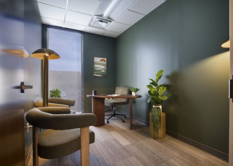 A small, modern office with green walls, a wooden desk, chair, two armchairs, potted plants, a tall lamp, a window with a blind, and a landscape artwork on the wall. The room is tidy and well-lit.