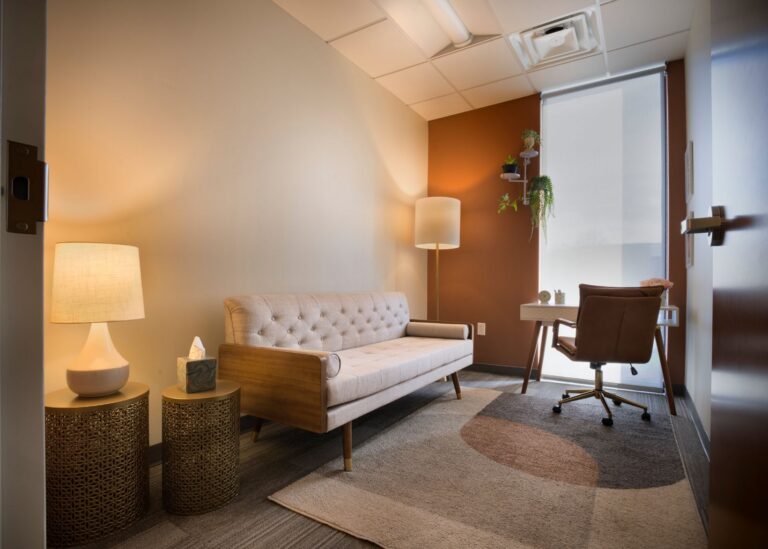 A cozy office with a beige sofa, two round side tables with lamps and tissues, a desk with a chair by a window, potted plants, and warm lighting. The decor is minimalist and inviting.