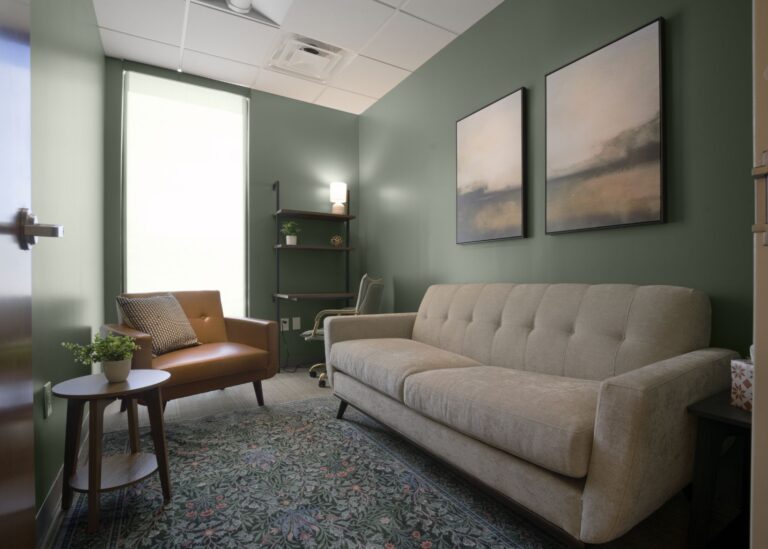 A small office with green walls, a beige sofa, a brown armchair, a side table with a plant, a patterned rug, a desk with a lamp, and two abstract paintings on the wall.
