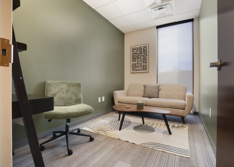 A modern office with green walls, a beige sofa, a green desk chair, a wooden coffee table, a small desk, an abstract rug, and a framed geometric art piece on the wall. Natural light comes through a tall window.