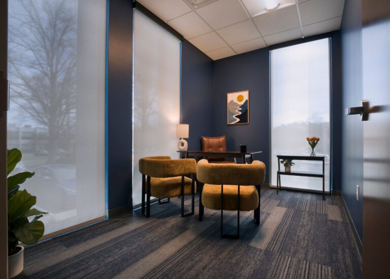Modern office with navy walls, two mustard chairs facing a desk with a leather chair, a small lamp, a framed abstract art piece, large windows with sheer blinds, a side table with flowers, and a potted plant in the corner.