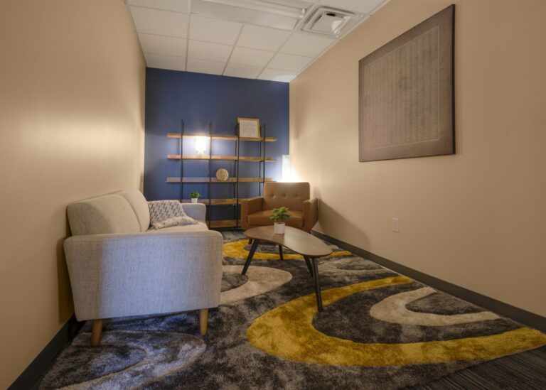A cozy, modern room with a beige sofa, brown armchair, small coffee table, abstract rug, wall shelf with decor, and soft lighting against a blue accent wall.