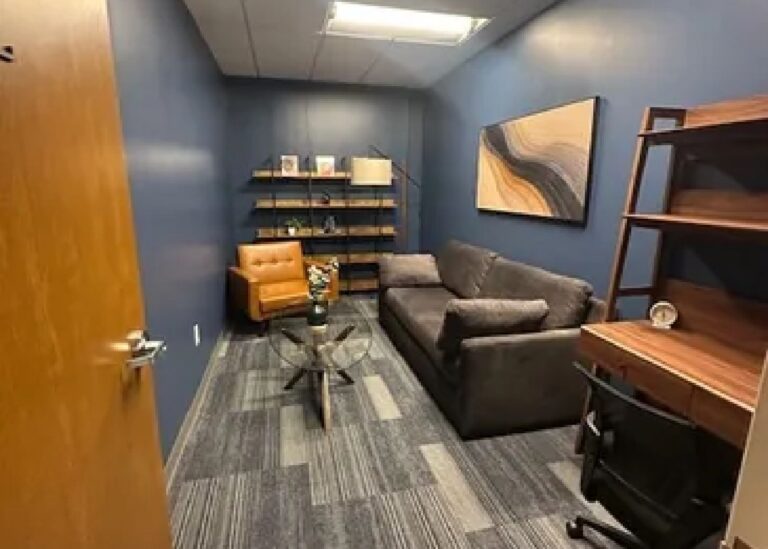 A small office with blue walls, a gray sofa, a brown leather chair, a glass coffee table, a wooden bookshelf with books and decor, a desk with a chair, and abstract wall art.