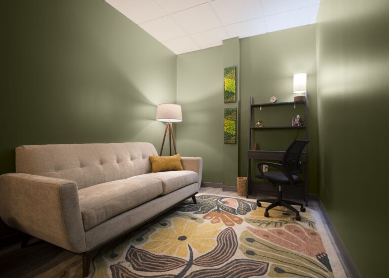 A cozy office with green walls, a beige sofa with a yellow pillow, a standing lamp, a patterned rug, and a black desk with a chair, lamp, and decorative shelves.